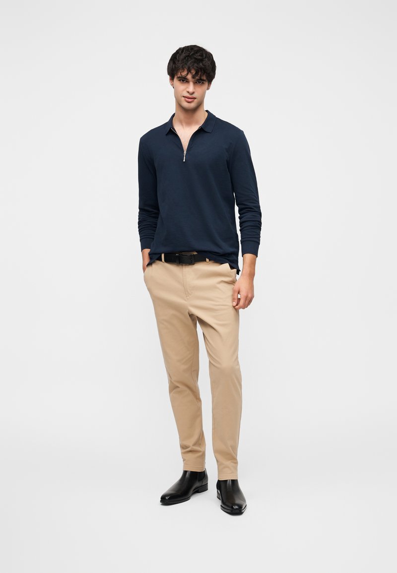 Marineblå langærmet polo med lynlåslukning ved kraven, kombineret med lyse beige bukser og sorte ankelstøvler. Enkel, moderne pasform med rene linjer.