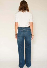 Jean évasé taille haute en denim bleu foncé, avec poches arrière, un design épuré et une texture légèrement usée, associé à une chemise blanche.