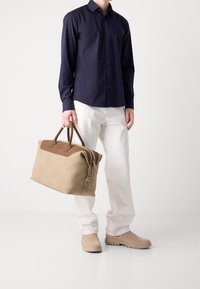 Beige leren sporttas met donkerbruine leren accenten, dubbele handvatten aan de bovenkant en ritssluiting. Beschikt over een gladde, zachte textuur.