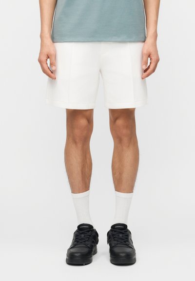 Homme portant un short blanc mi-cuisses, des chaussettes blanches montantes, des baskets noires et un t-shirt bleu clair, debout devant un fond blanc.