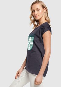 Marineblå bomulds T-shirt med korte ærmer og en afslappet pasform. Har et hvidt og grønt skjold-design med detaljerede udsmykninger på forsiden.