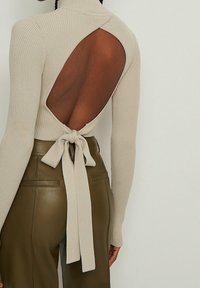Haut beige à côtes avec un col montant, présentant un dos ouvert et un nœud à la taille, associé à un pantalon en cuir olive foncé.