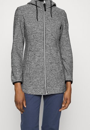 Persoon die een grijze hoodie met rits en zwarte koordjes draagt en marineblauwe broek, staand tegen een effen achtergrond.