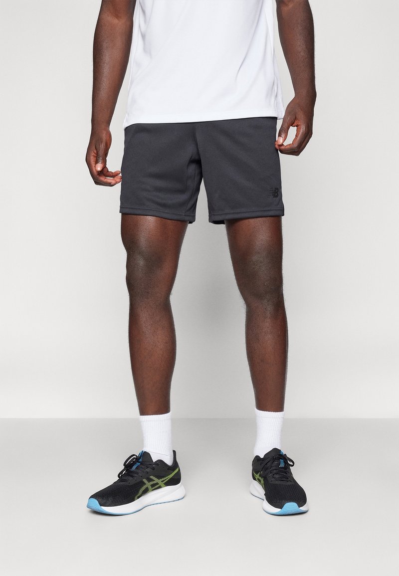 Man med svarta träningsshorts, vita ankelsockor, svarta löparskor med blå och gröna detaljer, och en vit t-shirt, som står på ett grått golv.