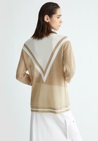 Maglione in rete beige con strisce diagonali bianche, caratterizzato da un design a V sulla schiena e orlo a coste. Tessuto leggero e testurizzato.