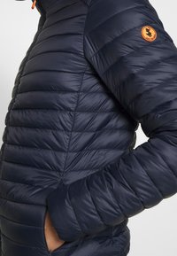 Marineblå dunjakke med quiltet design, der har en høj krave, lynlåslommer og et orange rundt logo på venstre arm.