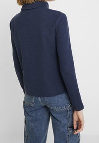 Maglione blu navy con colletto alto, realizzato in tessuto morbido. Presenta maniche lunghe e una vestibilità morbida, abbinato a pantaloni cargo azzurri chiari.