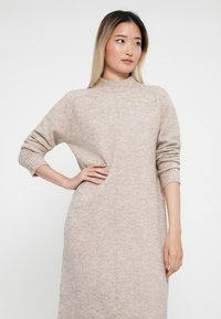 Vestito lavorato a maglia beige con collo alto falso, polsini a coste e silhouette dritta. Presenta una sottile texture melangiata e dettagli nelle cuciture laterali.