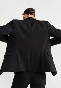 Černý blazer s přiléhavým střihem, s klopovými límci, strukturovanými rameny a bočními kapsami. Hladká tkanina bez viditelných vzorů.