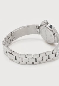 Ρολόι βραχιόλι από μέταλλο σε ασημί απόχρωση με κρύσταλλα Swarovski στο κούμπωμα και πίσω μέρος από ανοξείδωτο ατσάλι που φέρει το λογότυπο της μάρκας και την ένδειξη Swiss made.