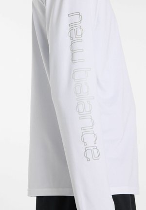 Camicia bianca a maniche lunghe con sottile logo di testo verticale sulla manica, abbinata a pantaloni neri, vista di profilo.