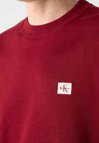 Gros plan d'une personne portant un sweat-shirt rouge Calvin Klein avec un petit patch logo blanc sur la poitrine.