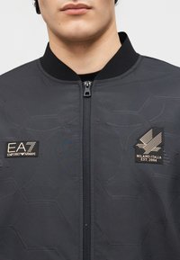 Άνδρας που φοράει σκούρο γκρι μπουφάν με φερμουάρ, με πατς EA7 Emporio Armani και Milano-Italia Est. 2004 στο στήθος και εξαγωνικό σχέδιο.