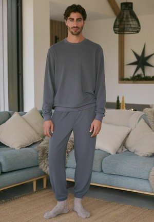 Grau geripptes Loungewear-Set mit einem Langarmoberteil und schmal zulaufenden Hosen, aus weichem Material gefertigt. Das Model steht auf einem strukturierten Teppich in der Nähe eines Sofas.