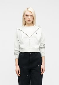 Γυναίκα με ανοιχτό ξανθά μαλλιά που φοράει μια ανοιχτό γκρι φούτερ με φερμουάρ πάνω από λευκή μπλούζα και μαύρο παντελόνι ψηλόμεσο, που στέκεται μπροστά σε απλό φόντο.