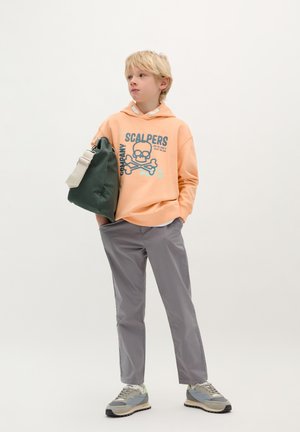 Sweat à capuche orange avec un graphisme de crâne, pantalon gris et baskets grises. Le mannequin porte un sac vert. L'arrière-plan est blanc uni.