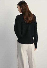 Kobieta z długimi brązowymi włosami w czarnym swetrze i białych spodniach stoi na tle jednolitego jasnego tła, odwrócona plecami do aparatu.