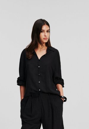 Camicia nera in lino con colletto, chiusure a bottoni e maniche arrotolate. Abbinata a pantaloni neri larghi coordinati.