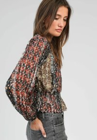 Femme portant un blouse à manches longues à motif floral avec une taille froncée et un jean gris, de profil contre un fond clair uni.