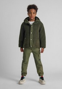 Giacca in pile verde con cappuccio, caratterizzata da una finitura testurizzata e bottoni a scatto. Abbinata a pantaloni cargo oliva e sneakers con dettagli colorati.