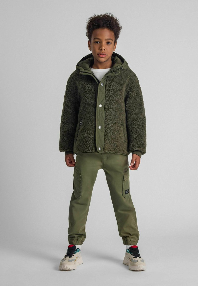 Giacca in pile verde con cappuccio, caratterizzata da una finitura testurizzata e bottoni a scatto. Abbinata a pantaloni cargo oliva e sneakers con dettagli colorati.