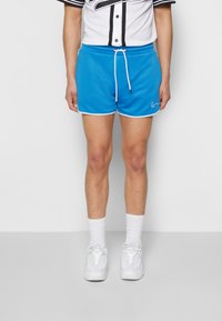 Blå idrettsshorts med hvit piping, elastisk midje og snor. Båret med en hvit kortermet skjorte og hvite joggesko.