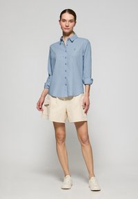 Camisa de botones azul claro con mangas enrolladas, combinada con pantalones cortos de cargo beige. La camisa presenta un sutil logo en el pecho.