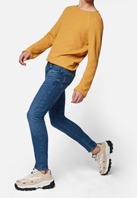 Suéter mostaza, vaqueros ajustados azules y zapatillas beige y negras. El suéter tiene mangas largas y un cuello redondo; los vaqueros son ajustados.