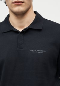 Gros plan sur une personne portant un polo noir Armani Exchange avec le texte du logo "ARMANI EXCHANGE MILANO / NEW YORK" sur la poitrine.