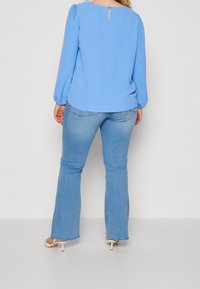 Blusa azzurro chiaro a maniche lunghe con maniche leggermente a sbuffo e dettaglio a chiave sul retro, abbinata a jeans svasati in denim chiaro.