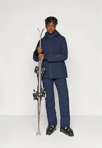 Άτομο με ναυτικό μπλε σκεπαστή ζακέτα, παντελόνια και μαύρες μπότες σκι που κρατάει τα σκι όρθια, φορώντας μαύρα γυαλιά σκι στο μέτωπο.