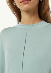 Hellblaues langärmliges Oberteil mit hohem Kragen, das über eine zentrale Naht und eine glatte Textur verfügt, aus einem weichen Stoff gefertigt.