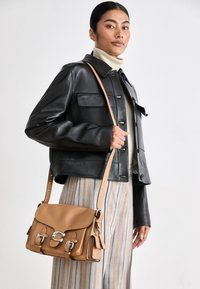 Tan leather crossbody bag with silver hardware, structured shape, buckle accents, and a detachable strap. Worn by a person in a black leather jacket.