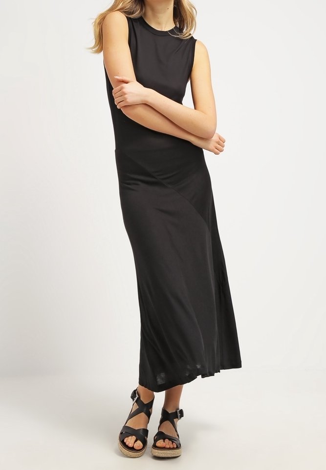 Femme debout, bras croisés, portant une robe longue noire sans manches et des sandales compensées noires avec brides à la cheville.