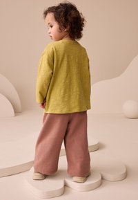 Maglia a maniche lunghe morbida e testurizzata di colore verde oliva abbinata a pantaloni rosa chiaro e larghi. Le calzature del bambino sono beige, posizionate su un piedistallo bianco a forma di nuvola.