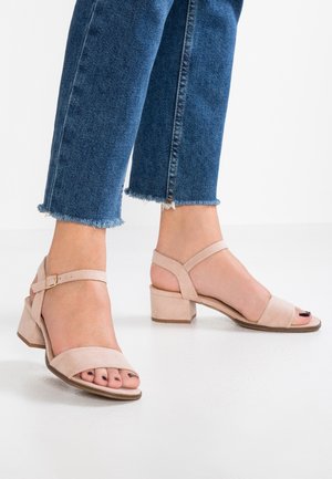 Fødder iført nude ruskindsklæder med blokhæl og ankelremme samt blå jeans med frynset kant mod hvid baggrund.