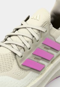 Beige träningsskor med en strukturerad ovandel, ljusgrå snören och tre framträdande rosa ränder på sidan. Gummisula och subtil logotyp.