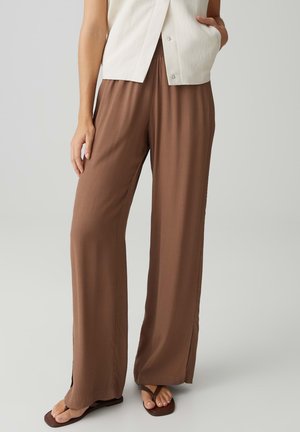 Donna che indossa pantaloni marroni larghi con spacchi laterali, top bianco senza maniche e sandali marroni, in piedi su uno sfondo chiaro e uniforme.