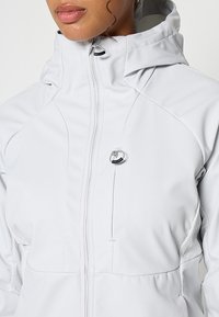 Jasnoszara kurtka zapinana na zamek, wykonana z gładkiego materiału, z kapturem, teksturowanymi bocznymi panelami i okrągłym akcentem z metalowego elementu z przodu.