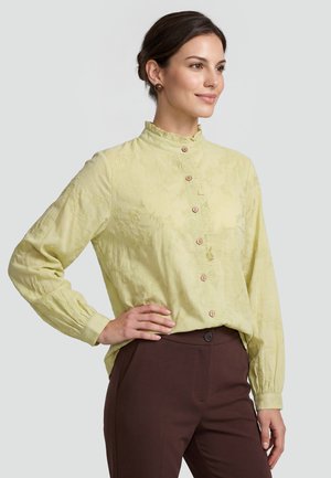 Frau trägt eine blassgelbe langärmelige Bluse mit Blumenstickerei und braune Hose, posiert mit einer Hand in der Hüfte.