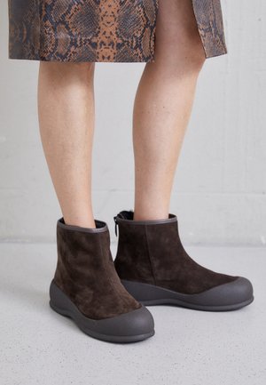 Jambes portant une jupe marron à imprimé serpent et des bottines en daim marron foncé avec des semelles épaisses en caoutchouc, sur un sol et un mur gris clair.