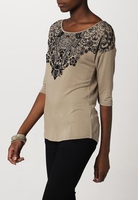 Beige top with black floral embroidery along the neckline, three-quarter sleeves, and a relaxed fit, paired with dark pants.