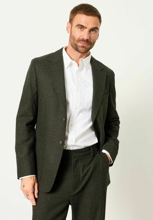 Mann steht, trägt einen dunkelgrünen Anzug mit weißem Hemd, eine Hand in der Tasche und auf der anderen Hand sichtbare Tattoos.