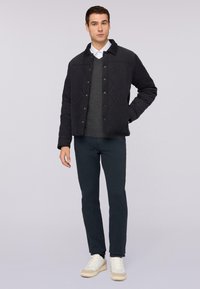 Zwart gewatteerd jack met een puntige kraag, drukknoopsluiting, grijze gebreide trui eronder en marineblauwe broek, gecombineerd met witte sneakers.