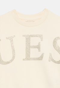 Cream-colored sweatshirt with a ribbed crew neck. Features large silver rhinestones forming the word "GUESS" across the front.