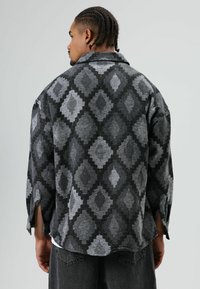 Man met gevlochten haar, gekleed in een grijs-zwarte jas met diamantpatroon, met de rug naar voren tegen een effen grijze achtergrond.