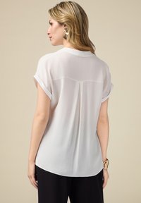 Blouse blanche à manches courtes avec un pli au dos et un ourlet arrondi. Tissu doux et léger avec des manches à revers. Associée à un pantalon noir.