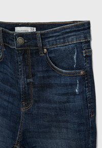 Jeans de mezclilla azul oscuro con corte recto, desvanecimiento sutil y detalles desgastados en la parte delantera. Accesorios en tono cobre y acentos cosidos.
