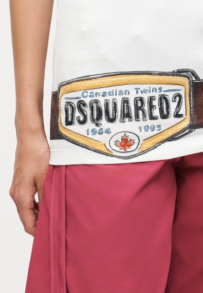 Κοντινό πλάνο ενός ατόμου που φοράει λευκό πουκάμισο με ζώνη με το λογότυπο DSQUARED2 και σκούρο ροζ παντελόνι.