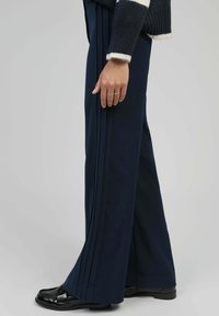 Pantalons larges bleu marine avec des plis verticaux, fabriqués en matière lisse. Associés à des chaussures noires polies, présentant une finition brillante.